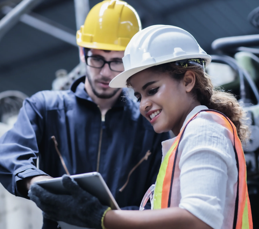 Two workers wearing hard hats and safety vests review information on a tablet in an industrial setting.